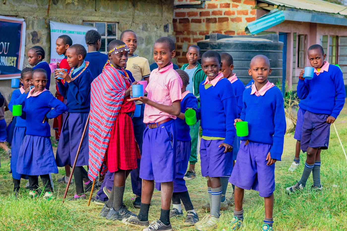 Children in school uniforms holding cups beside a water tank