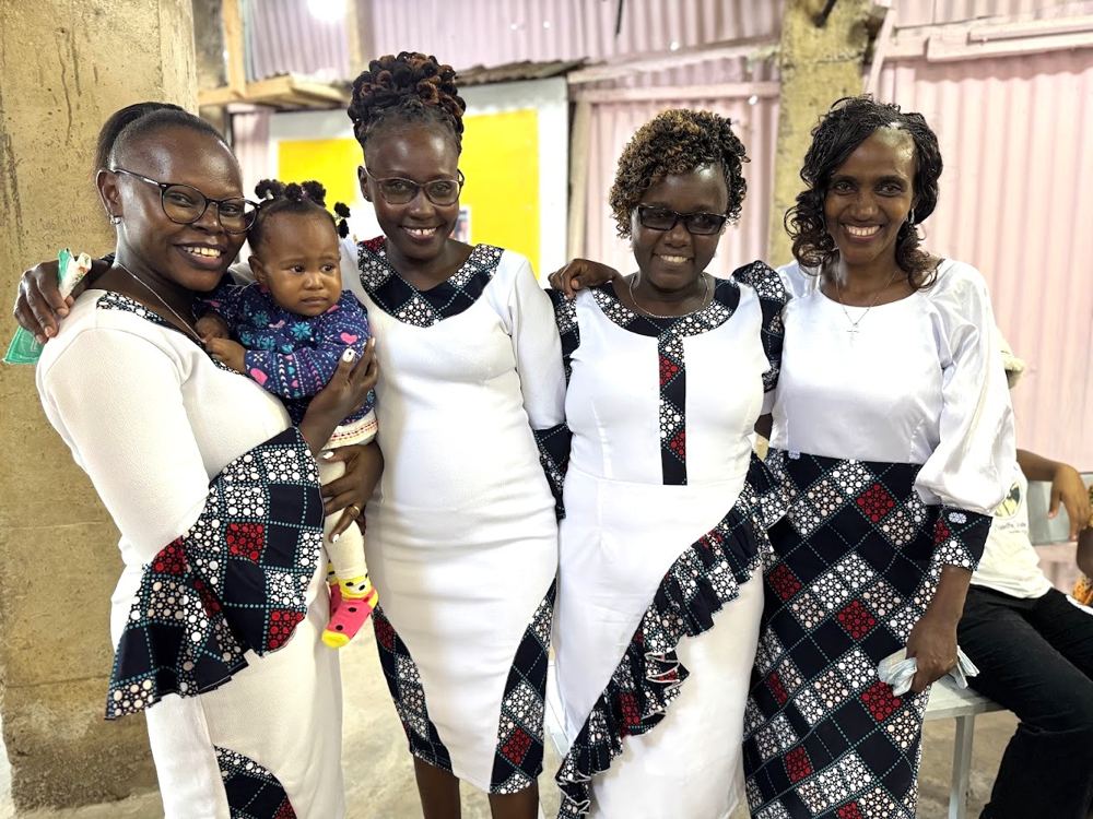 Four women in matching white dresses, one holding a baby, smiling together