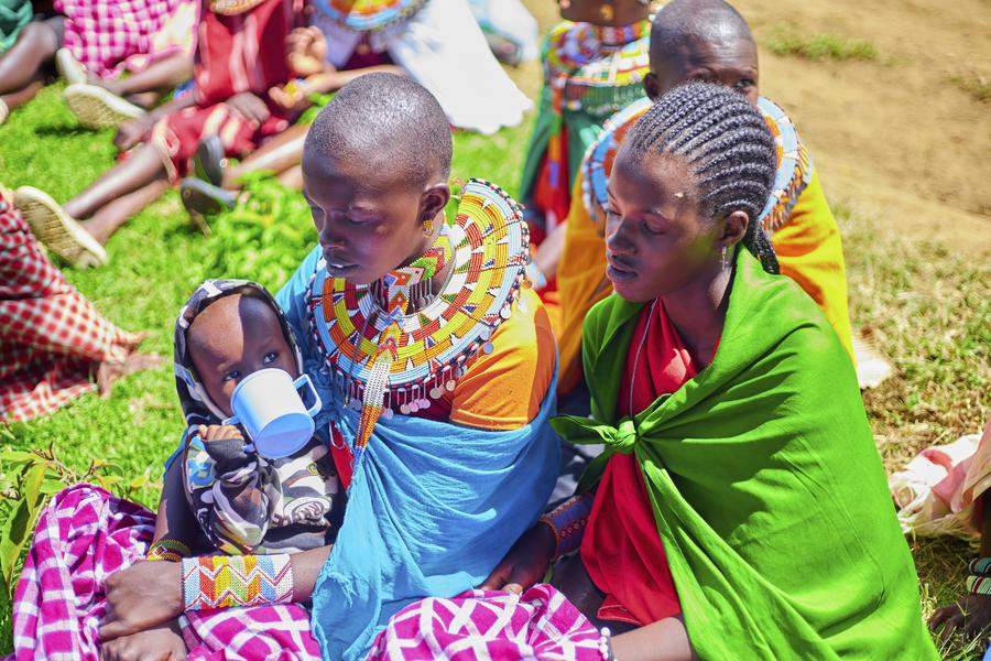 Samburu mother with infant drinking from cup, with friend beside her in beaded jewelry and orange shawl — GDP gathering, Northern Kenya