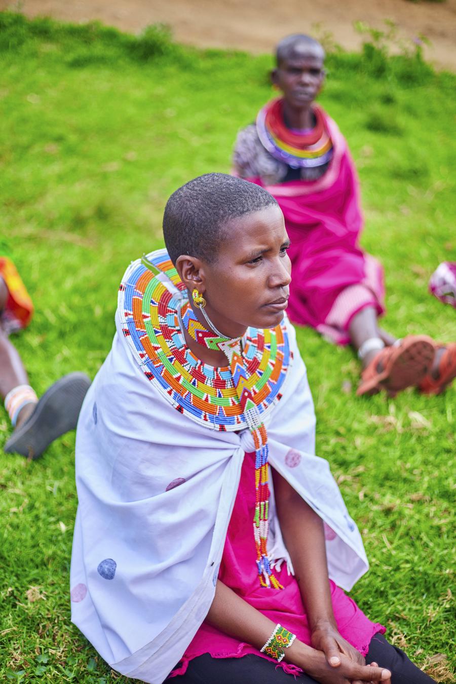Samburu woman portrait — white shawl, colorful beaded necklace, pink dress, seated on grass at GDP event, Northern Kenya