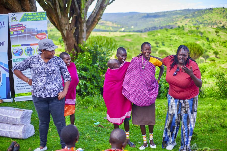 GDP team member with Samburu women and baby outdoors — St Rita school banner visible, smiling and joyful, Northern Kenya 2026