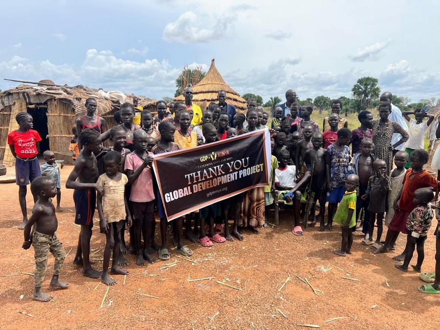 Children and youth near traditional huts holding GDP Kenya Thank You banner — humanitarian relief, East Africa