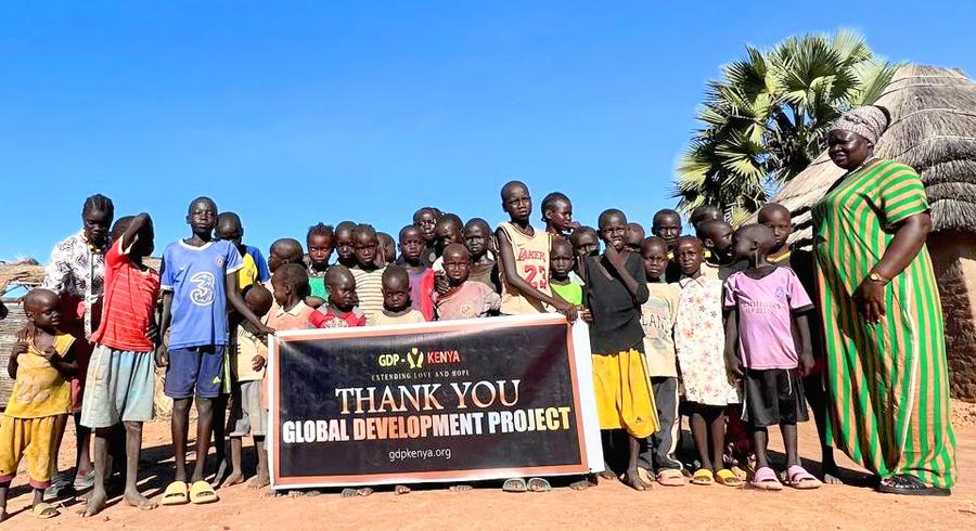 Village children and caregiver holding GDP Kenya Thank You banner in front of traditional thatched huts — humanitarian aid, East Africa